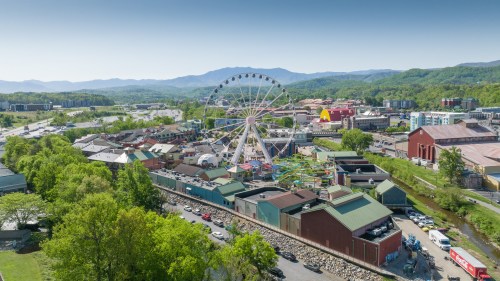 Aerial view of a vibrant amusement park with a Ferris wheel, surrounded by green hills and buildings.
