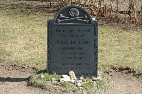 A gravestone with a skull and crossbones, honoring John Belushi, with dates and an inscription about his legacy.