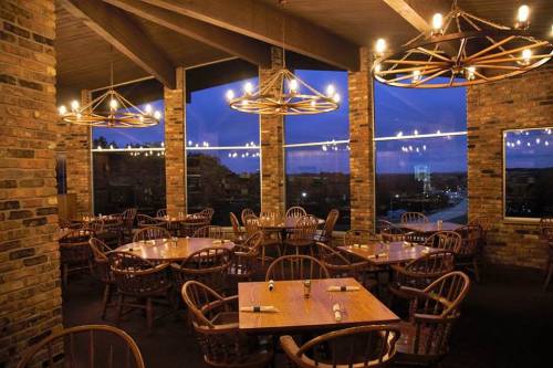 Cozy restaurant interior with wooden tables, chairs, and large windows, illuminated by warm hanging lights at dusk.