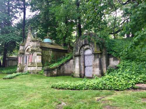 Overgrown stone mausoleums surrounded by lush greenery in a serene cemetery setting.