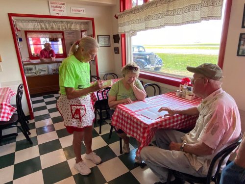A waitress in a green shirt takes an order from a couple at a checkered table in a cozy diner.