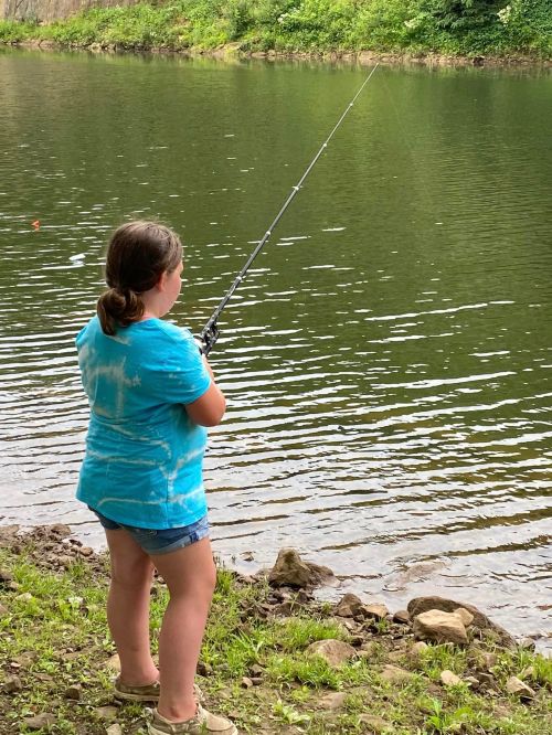 A child in a blue shirt stands by a calm lake, fishing with a rod, surrounded by greenery.