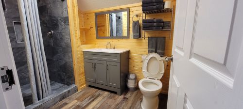 A modern bathroom featuring a shower, sink with cabinet, toilet, and wooden walls. Towels are neatly arranged on shelves.