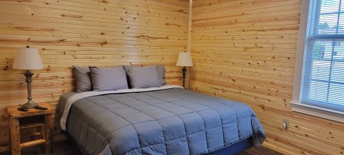 Cozy bedroom with wooden walls, a bed with gray bedding, and two lamps on nightstands. Natural light from a window.