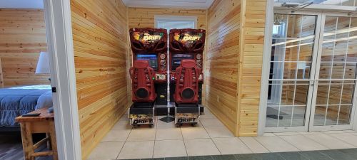 Two red arcade racing machines are positioned against a wooden wall in a room with tiled flooring.