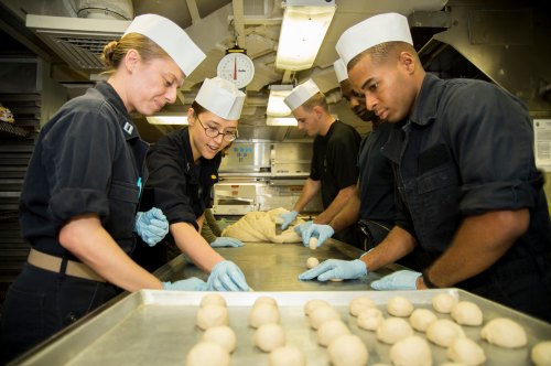 A group of individuals in chef hats and gloves prepare dough in a kitchen setting, focused on their task.