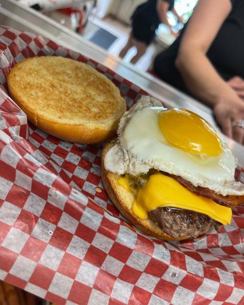 A burger with a fried egg, cheese, and bacon on a toasted bun, served on a red and white checkered paper.