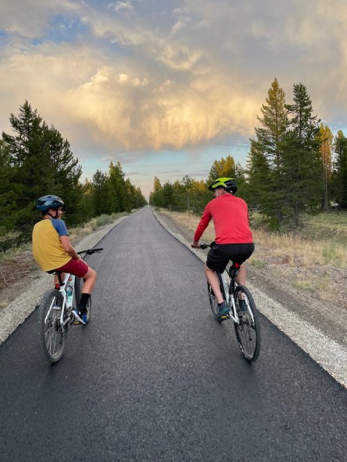 Two cyclists ride on a paved path through a forest, with a dramatic sky and clouds in the background.