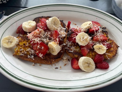 A plate of French toast topped with sliced bananas, strawberries, raspberries, and shredded coconut.
