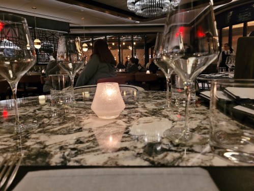 A dimly lit restaurant table set with wine glasses and a glowing candle on a marble surface, with diners in the background.