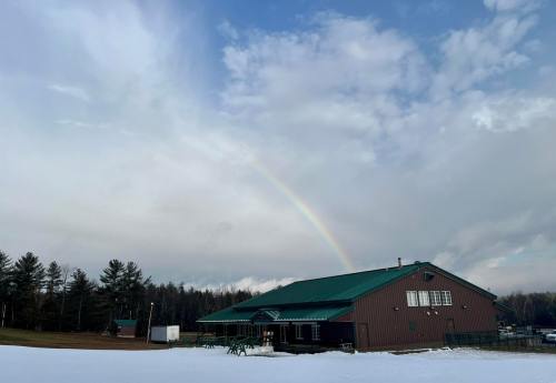A brown building with a green roof under a cloudy sky, featuring a rainbow in the background and snow on the ground.