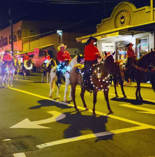 A nighttime parade featuring riders on horses adorned with lights, passing by shops in a festive atmosphere.