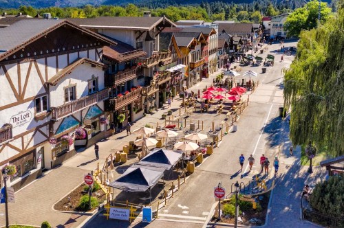 A charming street lined with shops and outdoor seating, featuring colorful umbrellas and a scenic backdrop of trees.