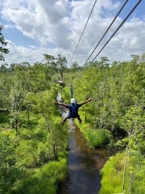 A person zip-lining over a lush green landscape with trees and a winding stream below.