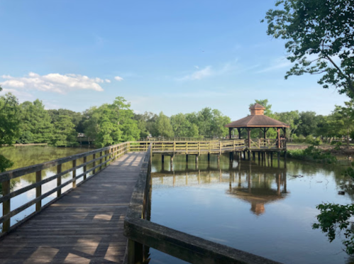 A wooden walkway leads to a gazebo by a calm lake, surrounded by lush greenery under a clear blue sky.