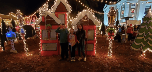 Three children pose in front of festive, illuminated holiday decorations, including decorated houses and trees.