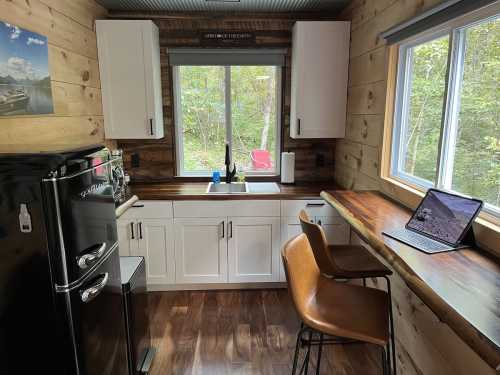 Cozy kitchen with wooden walls, a black fridge, white cabinets, and a small dining area by a window overlooking nature.