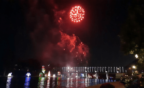 Colorful fireworks illuminate the night sky over a river, with festive lights reflecting on the water.