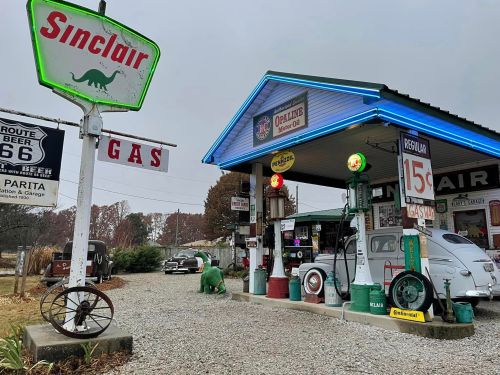 A vintage Sinclair gas station with retro pumps, classic cars, and a dinosaur statue, set against a cloudy sky.