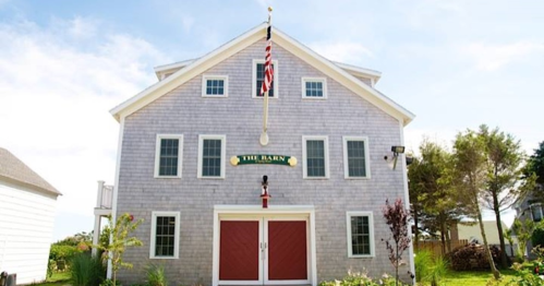 A large, gray barn with red doors, windows, and an American flag on top, surrounded by greenery.