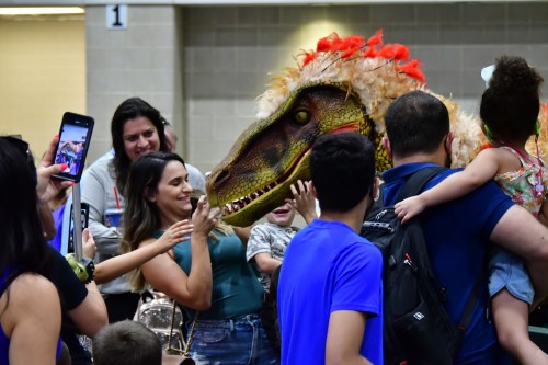 A crowd of people interacts with a large dinosaur costume at an event, with children reaching out to touch it.