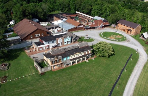 Aerial view of a rustic village with various buildings, green lawns, and trees in a rural setting.