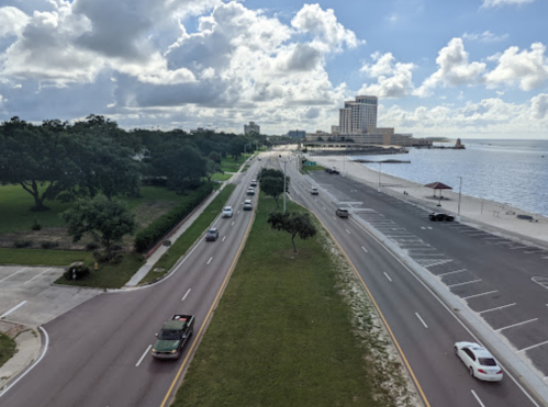 Aerial view of a road lined with trees, leading to a waterfront with buildings and a cloudy sky in the background.