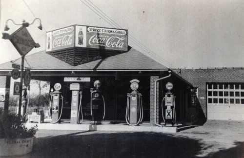 Black and white photo of a vintage gas station with Coca-Cola signage and old fuel pumps.