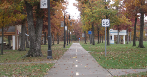 A tree-lined path with autumn foliage, featuring Route 66 signs and lampposts along the way.