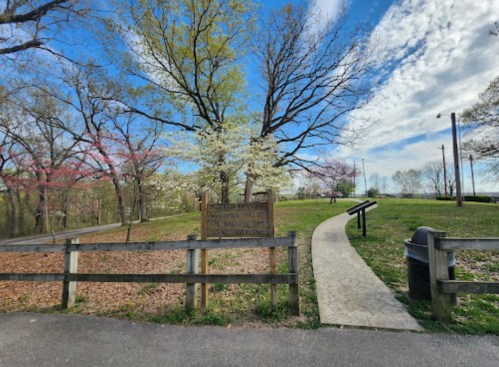 A park entrance with a wooden sign, trees in bloom, a winding path, and a trash can nearby under a blue sky.
