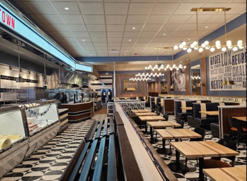 Modern restaurant interior with wooden tables, black chairs, and patterned flooring, featuring a counter and decorative lighting.