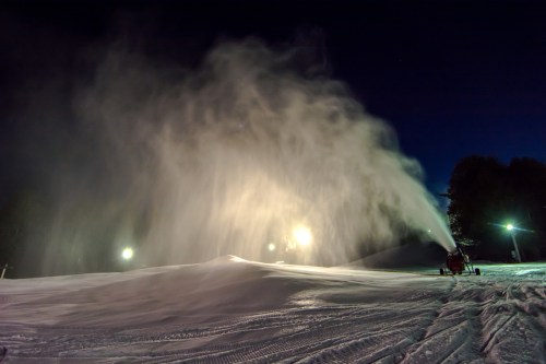 A snowmaking machine sprays snow onto a ski slope at night, illuminated by bright lights in the background.