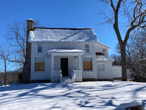 A white house with boarded windows, surrounded by snow and trees, under a clear blue sky.
