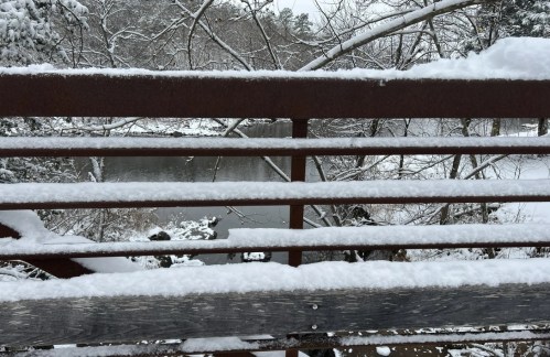 Snow-covered railing overlooking a serene, frozen river surrounded by trees in a winter landscape.