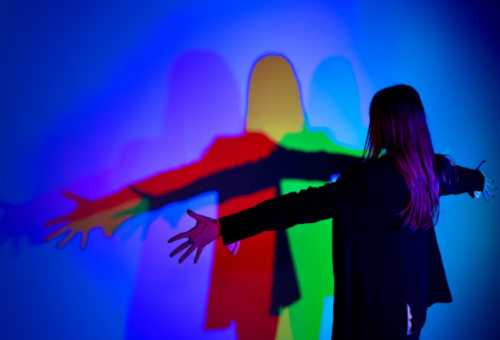 A person with outstretched arms casts colorful shadows against a blue background.