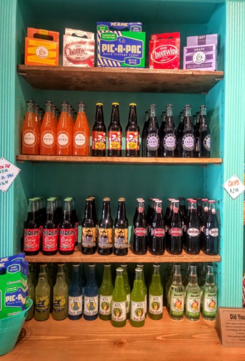 A colorful display of various soda bottles on wooden shelves, featuring brands like Cheerwine and other vintage-style drinks.