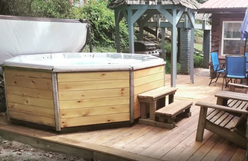 A wooden hot tub on a deck, surrounded by trees, with a gazebo and outdoor seating nearby.