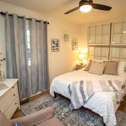 Cozy bedroom with a bed, decorative pillows, a chair, and light curtains, featuring a warm, inviting atmosphere.