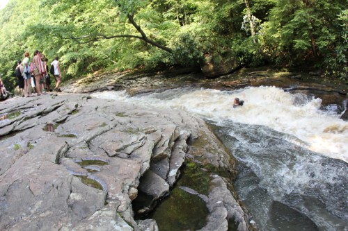 A group of people stands on rocky terrain by a rushing river, while one person swims in the turbulent water.