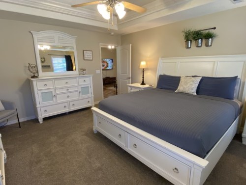 A cozy bedroom featuring a large white bed, dresser, and decorative plants on the wall. Soft lighting and carpeted floor.