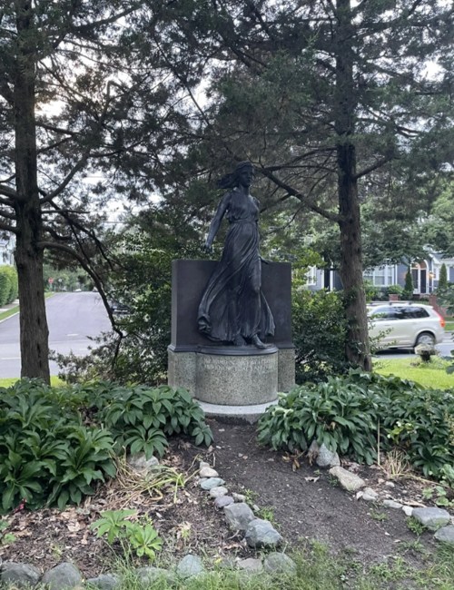 A bronze statue of a woman in a flowing dress stands on a stone pedestal, surrounded by greenery and trees.