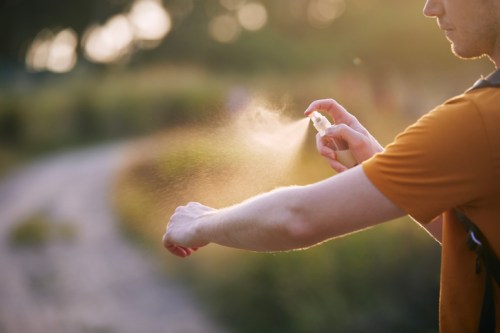A person in an orange shirt sprays insect repellent on their arm while outdoors in a sunlit, natural setting.