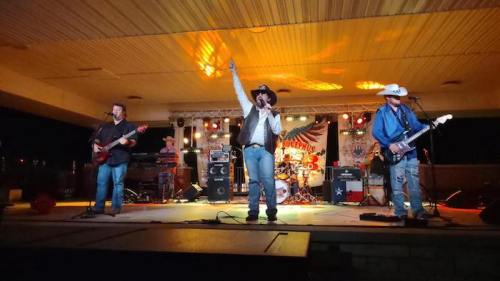 A country band performs on stage at night, with a singer raising his hand and musicians playing guitar and bass.