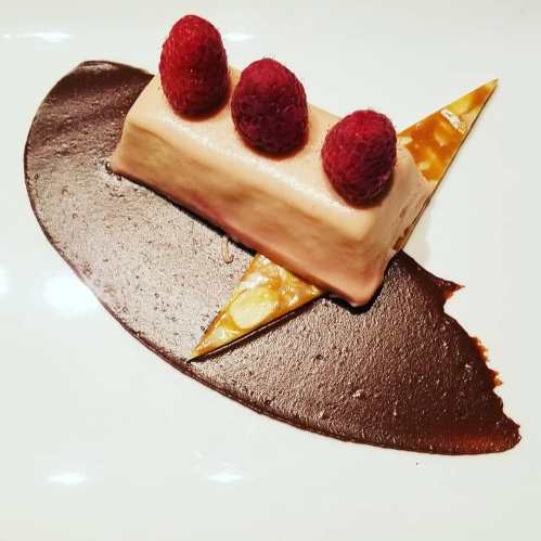 A dessert featuring a creamy mousse topped with raspberries, served on a chocolate sauce with a decorative caramel shard.