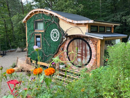 A whimsical tiny house with a green, vine-covered exterior and a round door, surrounded by colorful flowers and greenery.