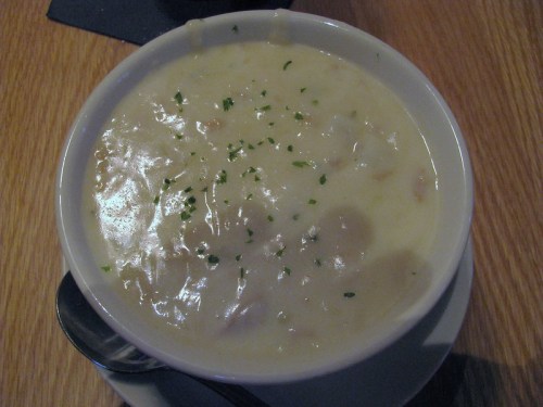 A bowl of creamy soup garnished with parsley, sitting on a wooden table with a spoon beside it.