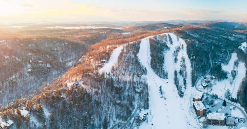 Aerial view of a snowy mountain landscape with ski slopes and a resort nestled among trees at sunset.