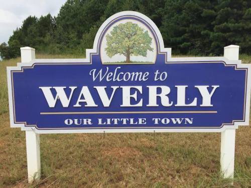 A blue and white welcome sign for Waverly, stating "Welcome to Waverly, Our Little Town," surrounded by greenery.