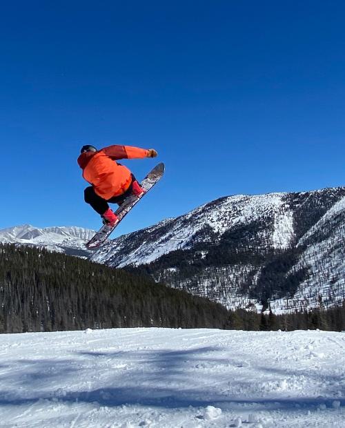 A snowboarder in an orange jacket jumps high above a snowy landscape with mountains in the background.