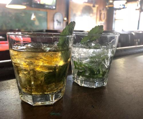 Two glasses on a bar: one with a golden drink and mint, the other with a clear drink and mint, both on a wooden surface.
